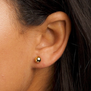 May include: A close-up of a person's ear with a small, round, gold earring. The earring is a simple, classic design.