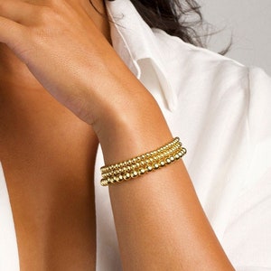May include: A set of four gold beaded bracelets stacked on a person's wrist. The bracelets are made of small, round beads.