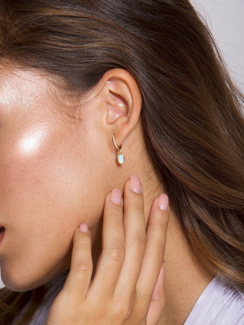 May include: Gold hoop earrings with a small oval opal dangle. The earrings are set with small clear stones.