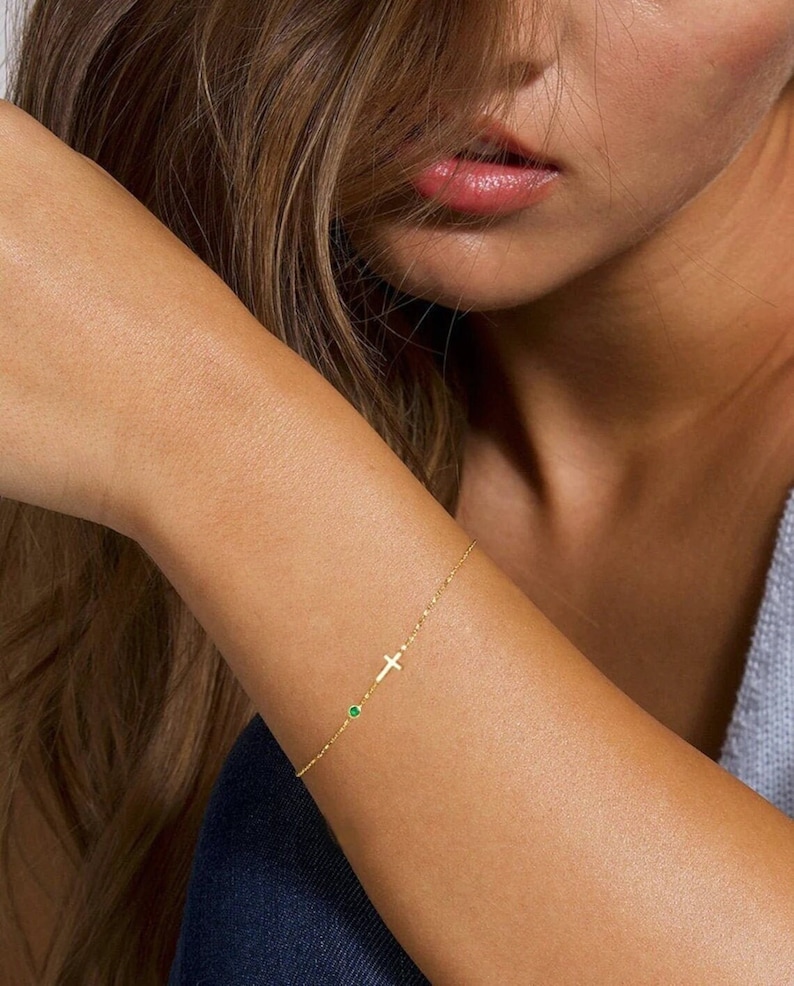 May include: A delicate gold chain bracelet featuring a small cross charm and a green gemstone accent.