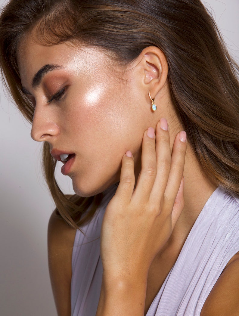 May include: A woman with long brown hair wearing a light purple sleeveless top and a gold hoop earring with a light blue gemstone pendant.