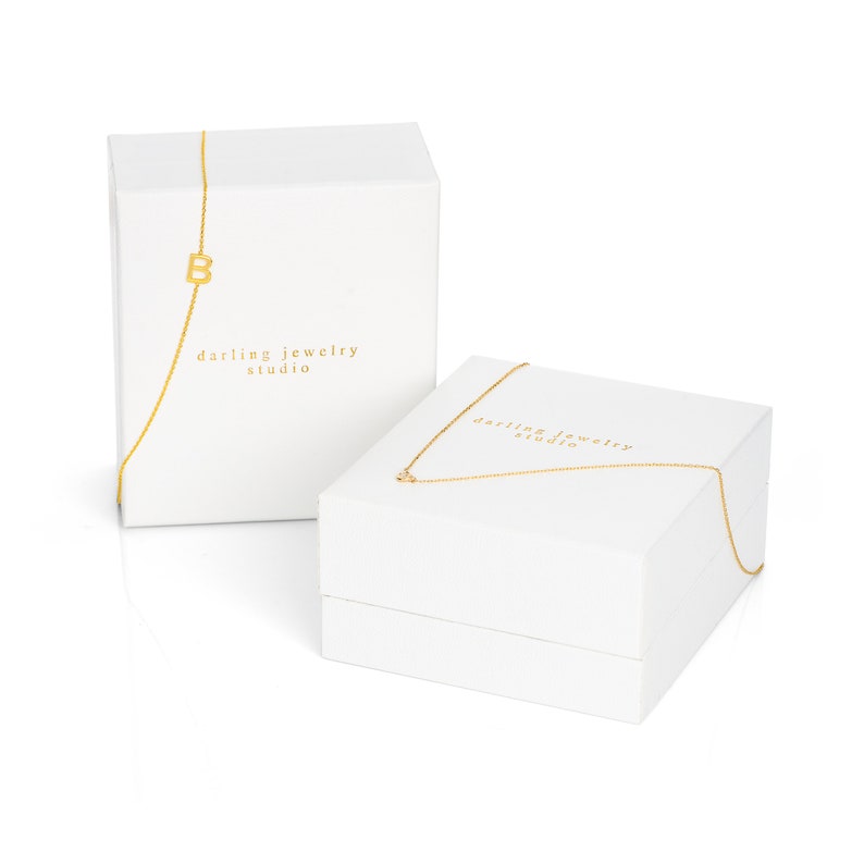 May include: Two white gift boxes with gold lettering. The top box has a gold chain with a letter "B" charm. The boxes say "darling jewelry studio".