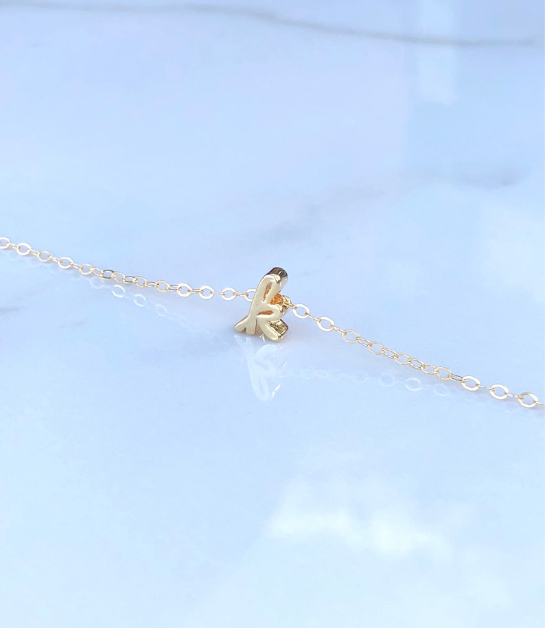 May include: A delicate gold chain necklace with a small, cursive letter 'K' pendant.