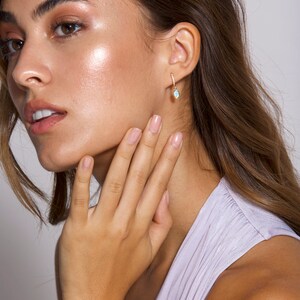 May include: A woman wearing a light purple top with a gold hoop earring with a light blue oval gemstone dangling from it.