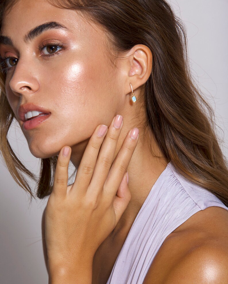May include: A woman wearing a light purple top with a gold hoop earring with a light blue oval gemstone dangling from it.