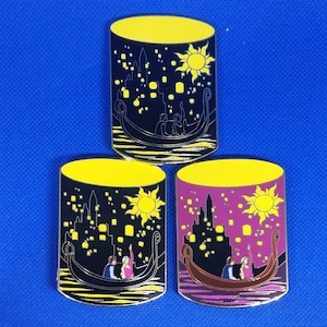 Könnte beinhalten: Drei Emaille-Pins, die eine Szene aus dem Disney-Film Rapunzel – Neu verföhnt zeigen. Jeder Pin zeigt eine Silhouette von Rapunzel und Flynn Rider in einem Boot mit Laternen, die im Himmel schweben. Die Pins sind in verschiedenen Farbschemata gehalten: Schwarz, Gelb und Lila.