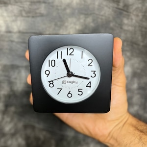 May include: A black square alarm clock with a white face and black hands. The clock face has black numbers and the word "bagby" printed in the center.