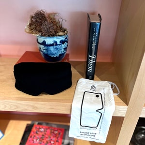 May include: A black sleep mask, a white drawstring bag with the text "bagby" and the words "Phone Free Bedrooms" printed on it, a book titled "Them" by Francine du Plessix Gray, and a blue and white ceramic bowl with dried plant material inside, all on a wooden shelf.