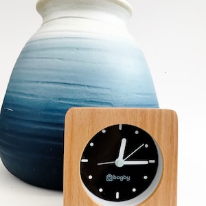 Peut inclure: Une horloge carr&eacute;e en bois avec un cadran noir et des aiguilles blanches. L'horloge a une bordure blanche et le mot "bagby" au centre. L'horloge est pos&eacute;e sur une surface blanche devant un vase en c&eacute;ramique bleu et blanc.