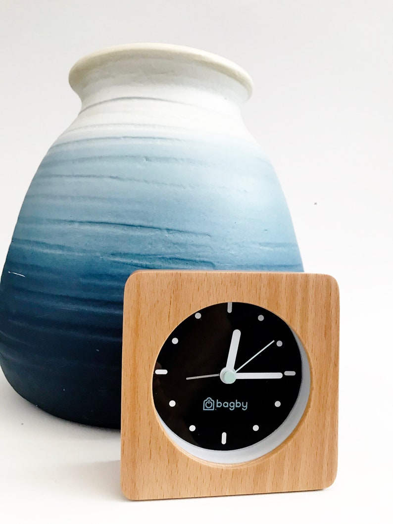 Peut inclure: Une horloge carr&eacute;e en bois avec un cadran noir et des aiguilles blanches. L'horloge a une bordure blanche et le mot "bagby" au centre. L'horloge est pos&eacute;e sur une surface blanche devant un vase en c&eacute;ramique bleu et blanc.