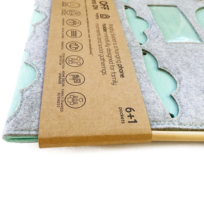 Op de afbeelding: Een grijze vilten telefoonhouder met een geschulpte rand en mintgroene vakken. De houder heeft een houten deuvel aan de onderkant en een kartonnen label met de tekst "Bagby Social" en "6+1 vakken".