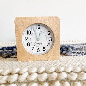 Puede incluir: Un reloj despertador cuadrado de madera con esfera blanca y números negros. La esfera tiene un segundero verde menta y la marca "bagby" impresa. El reloj está sobre una alfombra texturizada blanca y azul.