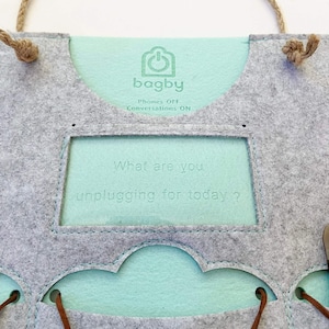 Op de afbeelding: Een grijze vilten tas met een lichtblauwe binnenkant en touwhandvatten. De tas heeft de tekst "bagby" en "Phones OFF Conversations ON". De zin "What are you unplugging for today?" is ook zichtbaar.