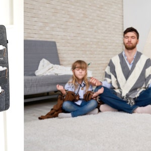 Op de afbeelding: Een donkergrijze vilten organizer met meerdere vakken met de labels "Marie", "Daddy", "Liona" en "Tablet". De organizer hangt aan een houten ladder. Op de achtergrond mediteert een gezin op een wit tapijt.