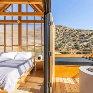 Peut inclure: Une cabane en bois avec une grande fen&ecirc;tre donnant sur un paysage d&eacute;sertique. La cabane a un lit avec des draps blancs et une petite table avec une horloge. Il y a une terrasse en bois &agrave; l'ext&eacute;rieur de la cabane avec un canap&eacute; noir et un foyer.