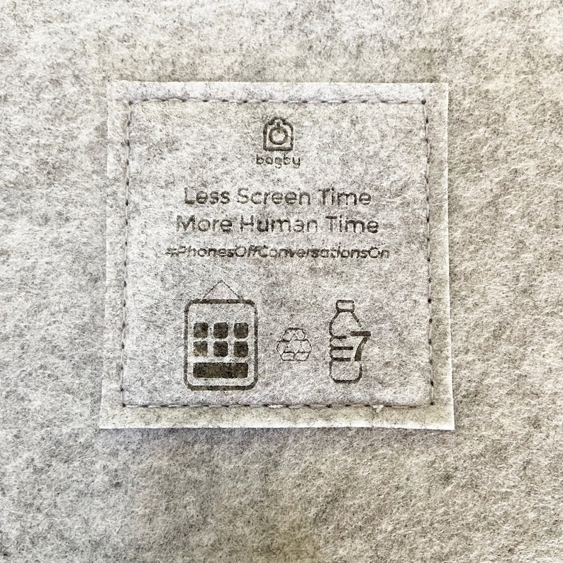 Op de afbeelding: Een grijs vilten vierkant met de tekst "Less Screen Time More Human Time" en de hashtag "#PhonesOffConversationsOn". Het vierkant heeft ook een telefoonpictogram, een recyclingsymbool en een flespictogram.