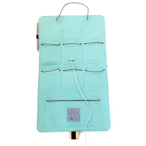 Op de afbeelding: Een lichtblauwe vilten organizer met meerdere vakken en een jute handvat. Het heeft een houten potlood en een wit snoer. De organizer is ontworpen om op te hangen en heeft een klein label aan de onderkant.