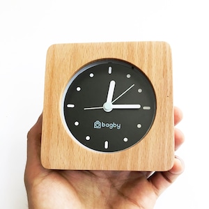 Peut inclure: Un r&eacute;veil en bois avec un cadran noir et des aiguilles blanches. L'horloge a un logo blanc qui dit "bagby" sur le cadran.