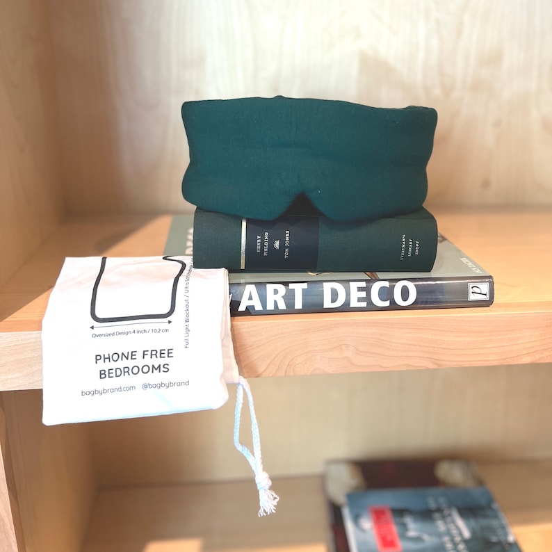 May include: A dark green sleep mask on top of two books. The top book is green and has the text "FIELDING" on the spine. The bottom book is a hardcover with the text "ART DECO" on the spine. A white drawstring bag with the text "PHONE FREE BEDROOMS" is in the foreground.