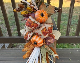 May include: A decorative autumn arrangement featuring two orange pumpkins, a burlap ribbon with a pumpkin print, dried wheat stalks, brown maple leaves, and eucalyptus sprigs. The arrangement is attached to a wooden lantern frame with vertical slats.