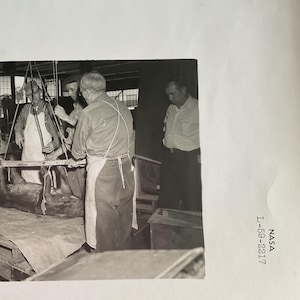 May include: A monochrome photograph showing a group of men in a workshop. They are attending to a suspended object, possibly a carcass, using ropes and pulleys. One man is wearing an apron. The image is labelled "NASA L-59-2217".