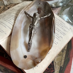 May include: A silver crucifix figure mounted inside a large, pearlescent seashell. The shell rests on open pages of an antique book with faded text. The crucifix is detailed, with arms outstretched.