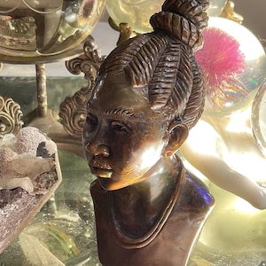 May include: A bronze-colored bust sculpture of a person with intricately carved hair styled in a bun. The sculpture features a detailed face with a necklace. The bust is displayed on a reflective surface, with decorative glass spheres in the background.