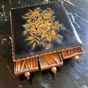 May include: A black matchbox with a gold floral design and three drawers filled with matches.