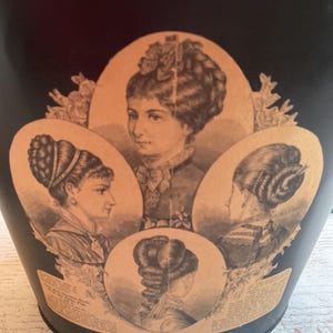 May include: A black cylindrical container featuring a beige graphic print of four oval portraits of individuals with intricate Victorian-style hairstyles. The design includes decorative floral elements and printed text surrounding the portraits.