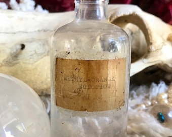 May include: A vintage glass bottle with a cork stopper and a label that reads "Methyl Orange Solution". The bottle is sitting on a white surface with a purple crystal and a white fluffy material.