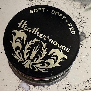 Könnte beinhalten: Eine schwarze runde Dose mit einem goldenen Design und dem Text "Soft Soft Red Heather Rouge" auf dem Deckel.