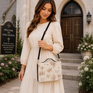 May include: A beige crossbody bag with a floral and cross design. The bag has a black strap and handle. The text on the bag includes the words "Grace", "Be Still and Know", and "Loved Beyond Measure". The image also shows a church sign that says "Welcome to Our Church".