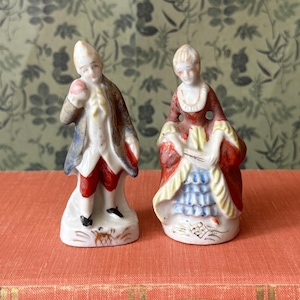 May include: Two porcelain figurines depicting a pair of individuals in historical attire. The male figure wears a blue coat and red breeches, while the female figure wears a red dress with a blue ruffled skirt. Both stand on white bases.