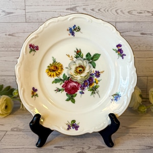 May include: A white ceramic plate with a scalloped edge and a gold rim. The plate features a floral design with yellow, red, purple, and white flowers. The plate is displayed on a black stand.