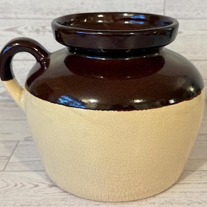 May include: A vintage stoneware jug with a dark brown top and handle, and a cream-colored base. The jug has a rounded body and a wide opening, with a capacity of approximately 1 gallon.