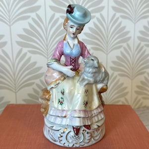 May include: A porcelain figurine of a person wearing a pink bodice and a cream-colored skirt with floral accents, seated on a base while holding a small gray lamb. The person wears a light blue hat with a dark red detail and has red shoes.