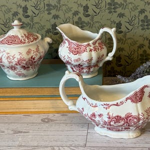 May include: A three-piece antique white porcelain serving set with a red floral pattern. The set includes a lidded sugar bowl, a creamer, and a gravy boat. The pieces are arranged on a stack of vintage books.