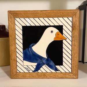 May include: Square framed artwork featuring a white goose with an orange beak and a blue scarf. The goose is set against a black background, framed by a white and black striped border, and a wooden frame. The artwork is signed in the bottom left corner.