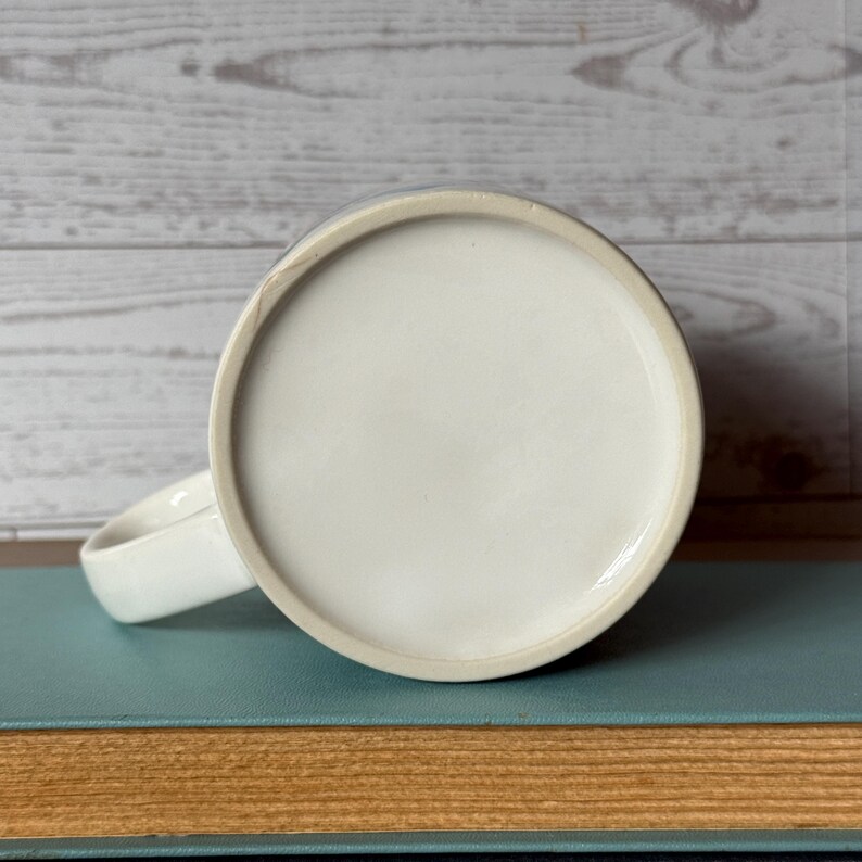 Puede incluir: Una taza de cer&aacute;mica blanca con base redondeada y asa curva. La taza est&aacute; sobre un libro azul, con un fondo de madera clara. El interior de la taza no es visible.