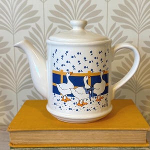 May include: A white ceramic teapot with a lid, featuring a graphic print of three white geese with orange beaks and feet. The geese are positioned in front of a blue and yellow fence, surrounded by small blue flowers on a white background.