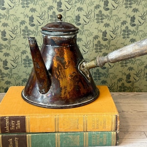 May include: Antique copper tea kettle with a wooden handle and lid. The kettle has a dark, aged patina and sits atop two vintage books with gold lettering. The background features a green floral wallpaper.