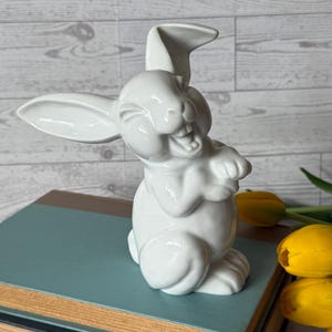 May include: A white ceramic bunny figurine with a smiling expression, upright ears, and paws clasped together. The bunny is positioned on a stack of books with yellow tulips in the background. The figurine is a decorative item.