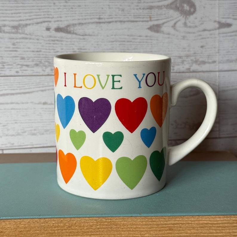 Puede incluir: Taza de cer&aacute;mica blanca con las palabras "I LOVE YOU" en colores arco&iacute;ris. La taza est&aacute; decorada con corazones coloridos en un patr&oacute;n de arco&iacute;ris. La taza tiene un asa curva.