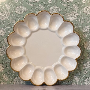 May include: A white ceramic oyster plate featuring a scalloped edge with a gold-painted rim. The plate has a central circular area surrounded by twelve individual oval-shaped indentations designed for holding oysters, with a glossy finish.