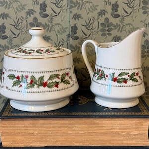 May include: A white porcelain sugar bowl with a lid and a matching creamer pitcher. Both pieces feature a decorative band of green holly leaves and red berries, accented with thin gold trim along the rims and base edges of the vessels.