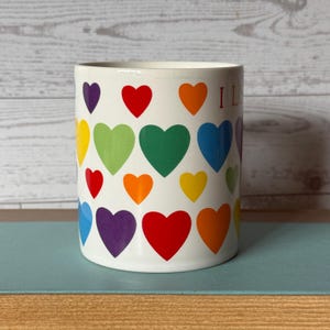 Puede incluir: Taza de cer&aacute;mica blanca decorada con corazones coloridos en un patr&oacute;n de arco&iacute;ris. La taza tiene forma cil&iacute;ndrica y la palabra "I L" es visible en el lateral. Los corazones son en tonos morado, rojo, naranja, amarillo, verde y azul.