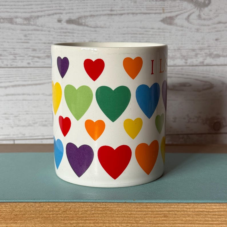 Puede incluir: Taza de cer&aacute;mica blanca decorada con corazones coloridos en un patr&oacute;n de arco&iacute;ris. La taza tiene forma cil&iacute;ndrica y la palabra "I L" es visible en el lateral. Los corazones son en tonos morado, rojo, naranja, amarillo, verde y azul.