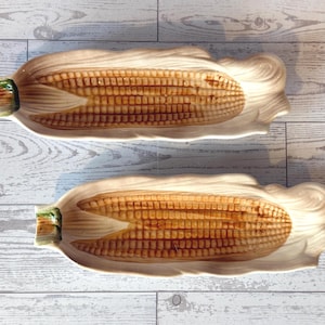 May include: Two ceramic corn-shaped serving dishes. The dishes are off-white with a detailed depiction of corn on the cob in shades of brown and yellow. The husks are sculpted to create a realistic look. The ends of the corn have green accents.