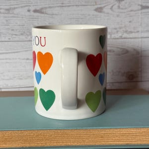 Puede incluir: Taza de cer&aacute;mica blanca con asa, decorada con corazones de colores rojo, naranja, verde y azul. La palabra "YOU" est&aacute; impresa en el lateral de la taza. La taza est&aacute; sobre una superficie azul.