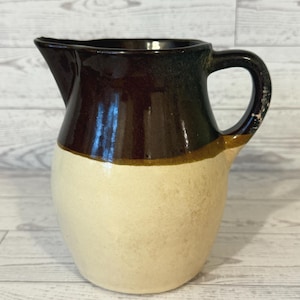 May include: A vintage ceramic pitcher with a cream-colored base and a dark brown top. The pitcher has a handle and a spout for pouring. A thin band of tan separates the two colors. The pitcher is likely used for serving beverages.